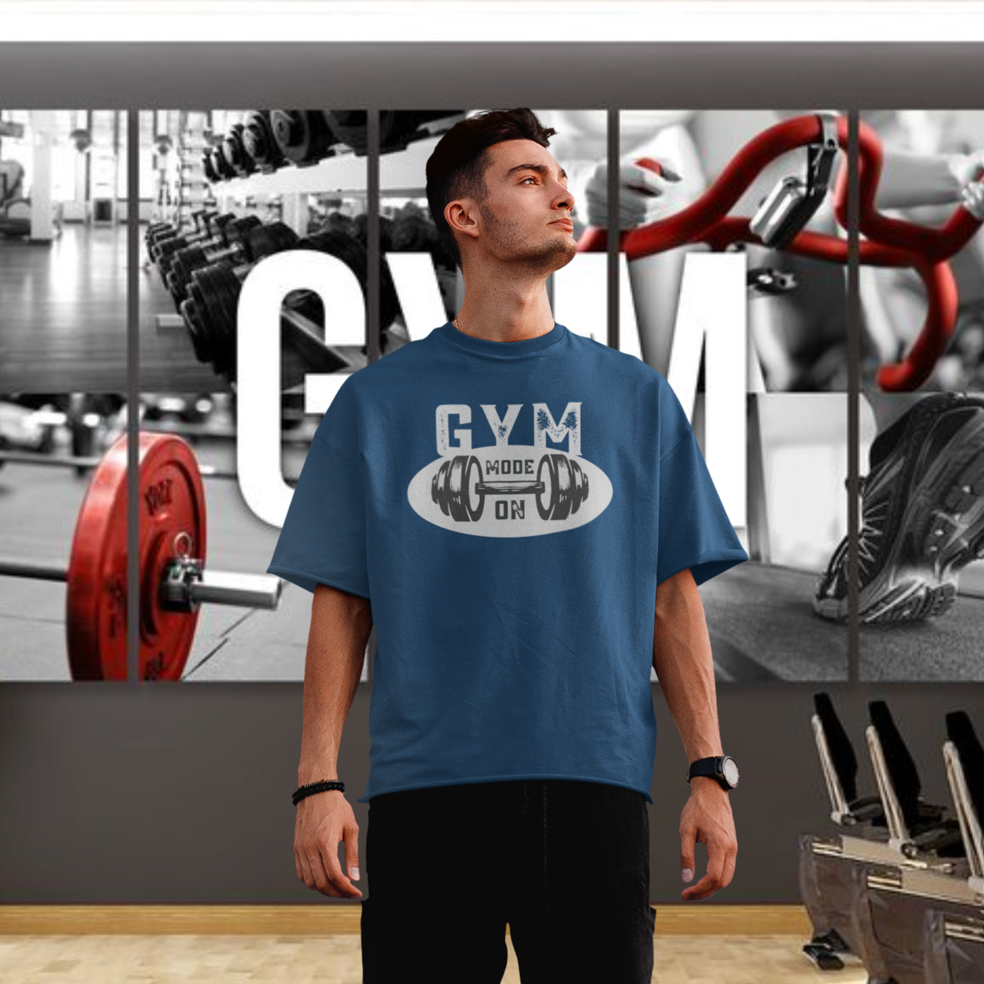 A man wearing a blue oversized round neck T-shirt with the text 'GYM' printed on the front, standing in a gym setting.